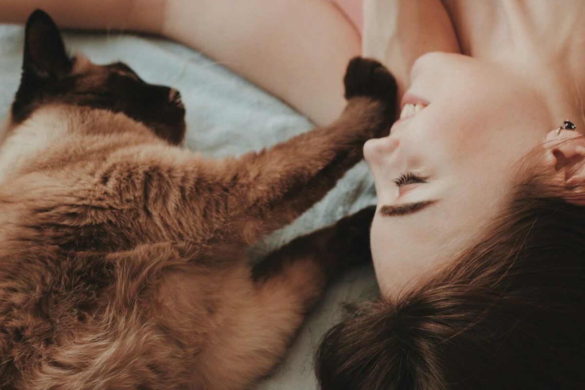 Woman laying next to brown cat with its paw on her face. 