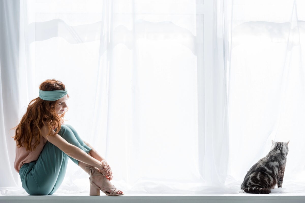 attractive young woman sitting on windowsill with cute tabby cat